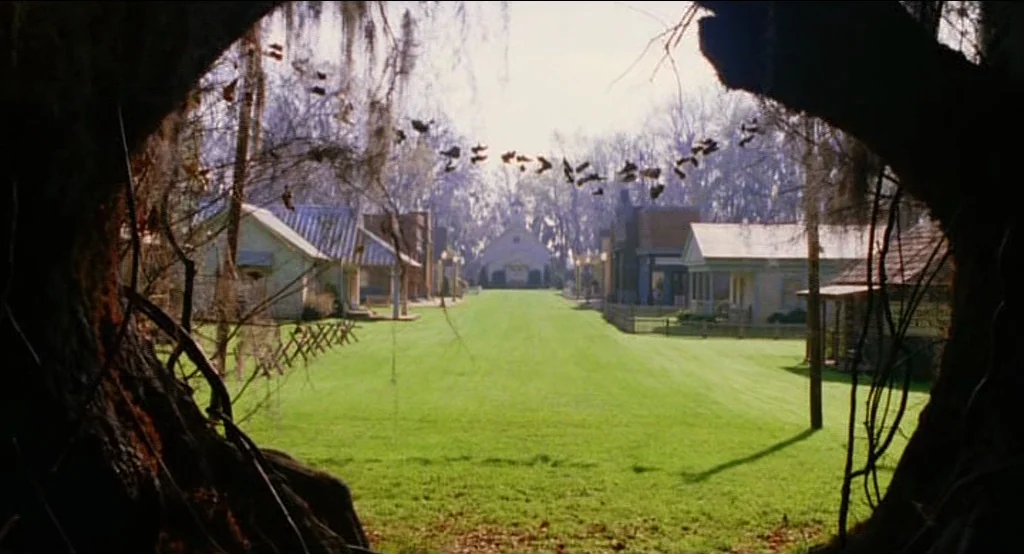 Big Fish (2003) movie still: establishing — A manicured green lawn leads toward a distant church building flanked by small houses. A line of shoes…