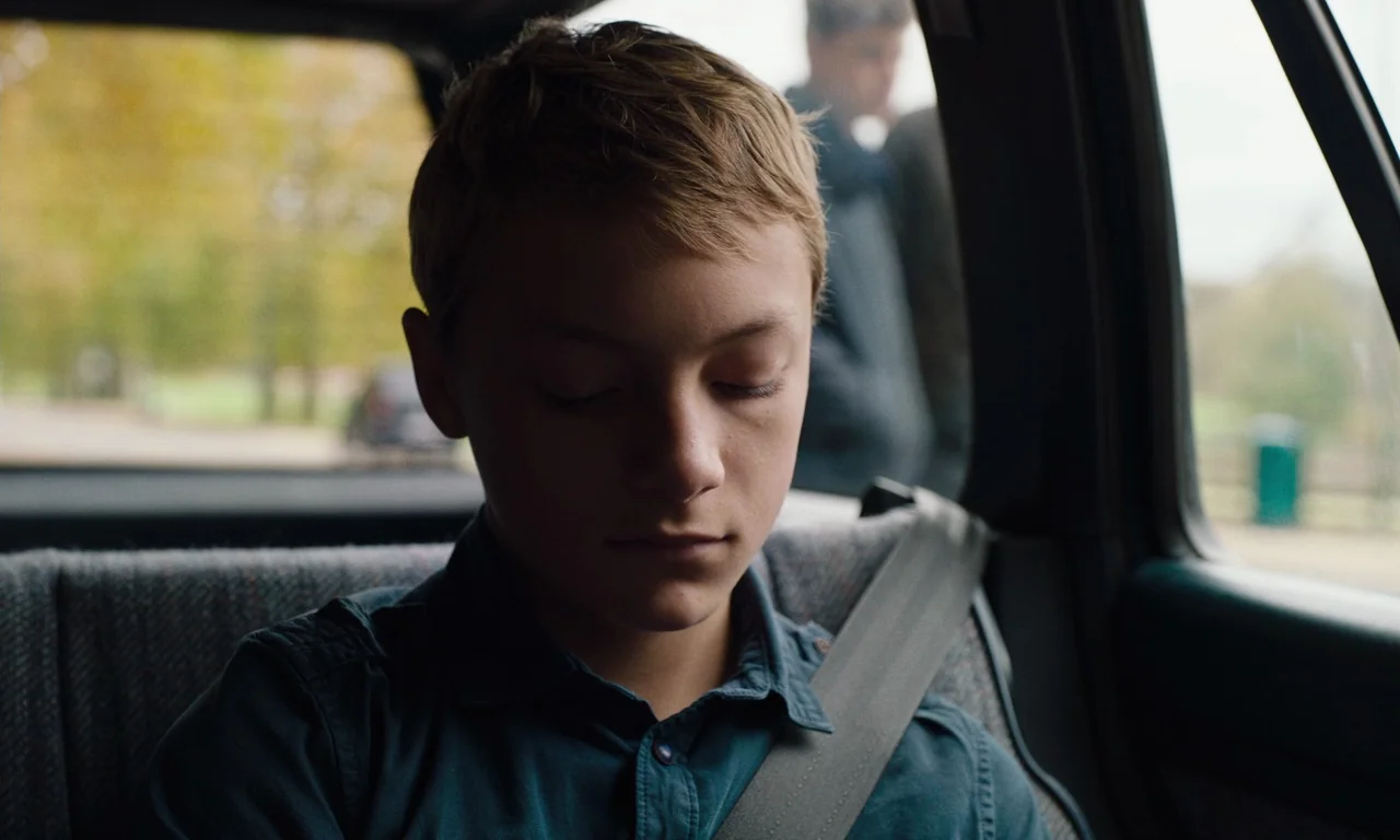 Close (2022) movie still: closeup — A teenage boy sits in the back of a car with his eyes closed. A blurred figure stands outside the window against…