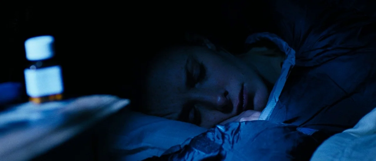 The Descent (2005) movie still: closeup — A dark bedroom illuminated by cool blue tones and deep shadows at night. A woman lies asleep on a pillow…