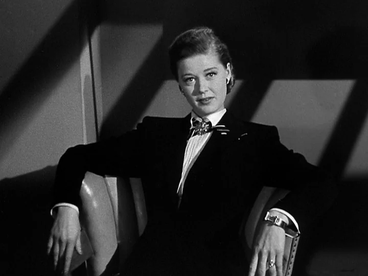 Nightmare Alley (1947) movie still: medium — A woman sits in a chair against a wall marked by angular shadows. She wears a tailored suit and sits…