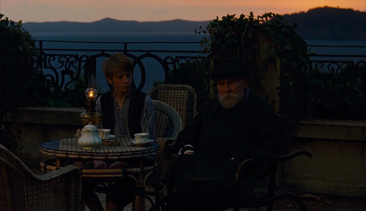 Renoir (2012) movie still: medium — A stone terrace overlooks the dark sea at dusk with distant rolling hills visible. A young boy and an elderly…