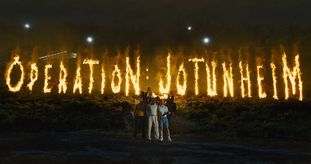 The Suicide Squad (2021) movie still: wide — A dark night landscape features large burning text reading Operation Jotunheim on a hillside. Three men…