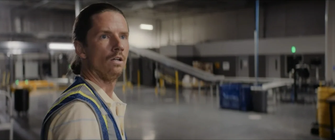The Forge (2024) movie still: medium — An industrial facility features conveyor belts and grey concrete flooring in a large room. A man stands in…