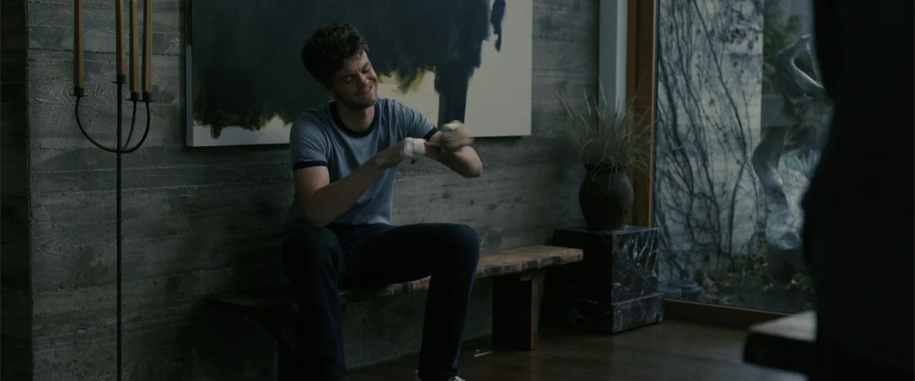 Companion (2025) movie still: medium — A modern room features a dark concrete wall with large abstract artwork and wood floors. A young man sits on…