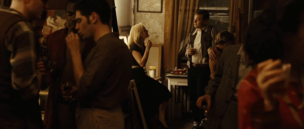 Revolutionary Road (2008) movie still: medium — A dimly lit, cluttered apartment room features several people standing in conversation at night. A…