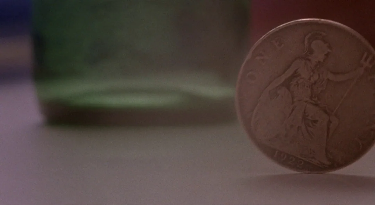 White Of The Eye (1987) movie still: closeup — A close-up view shows a 1922 British penny standing upright on a flat surface. A blurry, dark green…