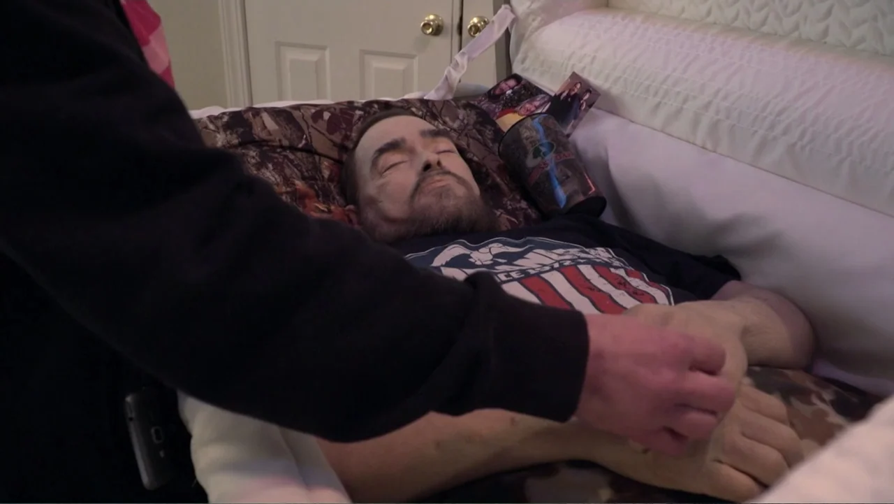 Meth Storm (2017) movie still: medium — A lined casket contains a man wearing a casual t-shirt on a camouflage pillow. A hand reaches into the frame…