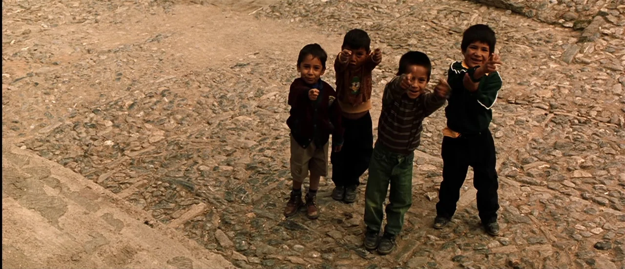The Mexican (2001) movie still: fullbody — Four children stand together on a textured cobblestone path surface. They gesture toward the camera lens…