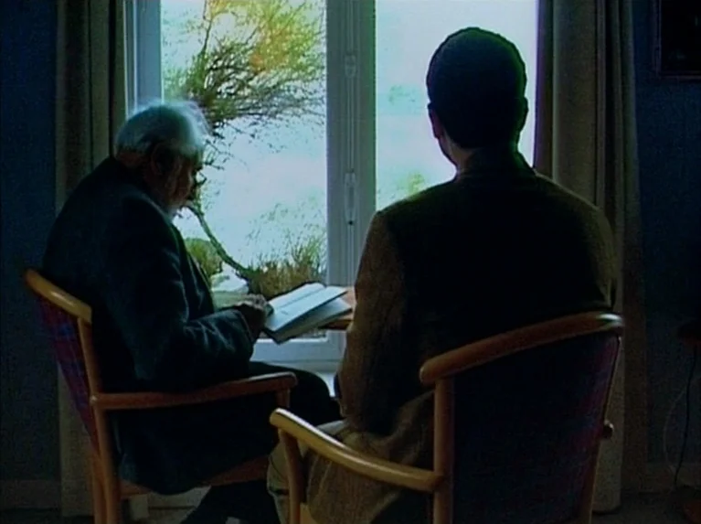 Eloge de L’amour (2001) movie still: medium — Two men sit in wooden armchairs facing each other by a brightly lit window. The older man reads from…