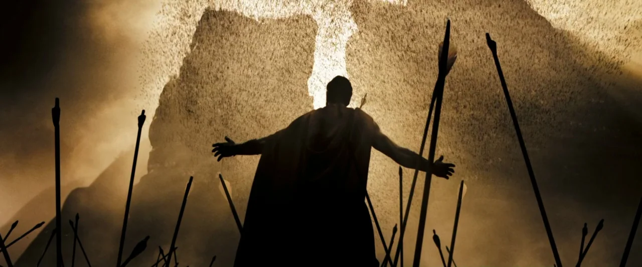 300 (2006) movie still: fullbody — A dust-filled battlefield shows an ancient warrior standing before a wall of incoming arrows. The man extends his…