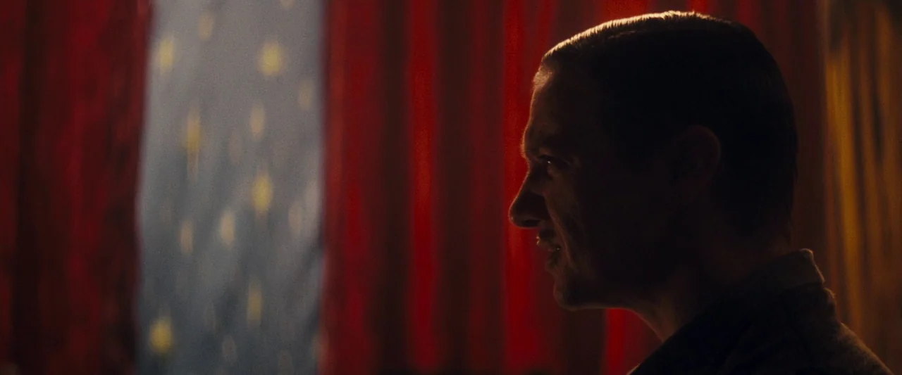 The Immigrant (2013) movie still: closeup — A dimly lit interior space is defined by deep red velvet curtains. A man is viewed in profile against…