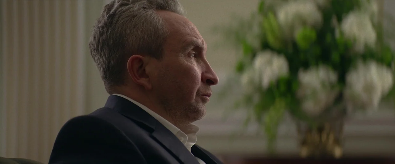 Fair Play (2023) movie still: closeup — An indoor office setting with a large floral display visible in the blurred background. A mature man in a…