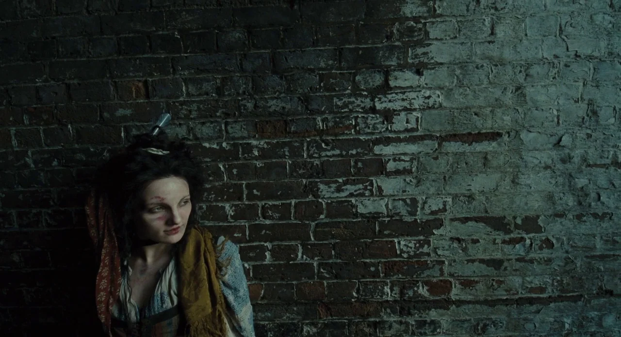 Les Misérables (2012) movie still: medium — A damp interior space features a rough-textured, dark, aged brick wall with areas of white paint. A…