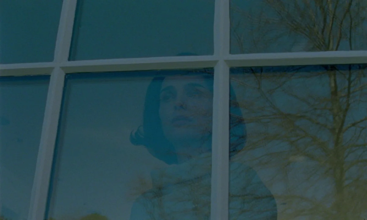 Jackie (2016) movie still: closeup — A woman appears through the reflection of a multi-paned window against a teal sky. She stands behind the glass…