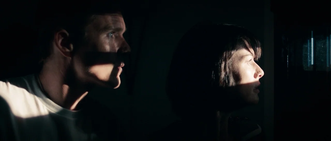 The Purge (2013) movie still: medium — Two individuals stand side by side in a darkened room, illuminated by a sliver of light. They look toward the…