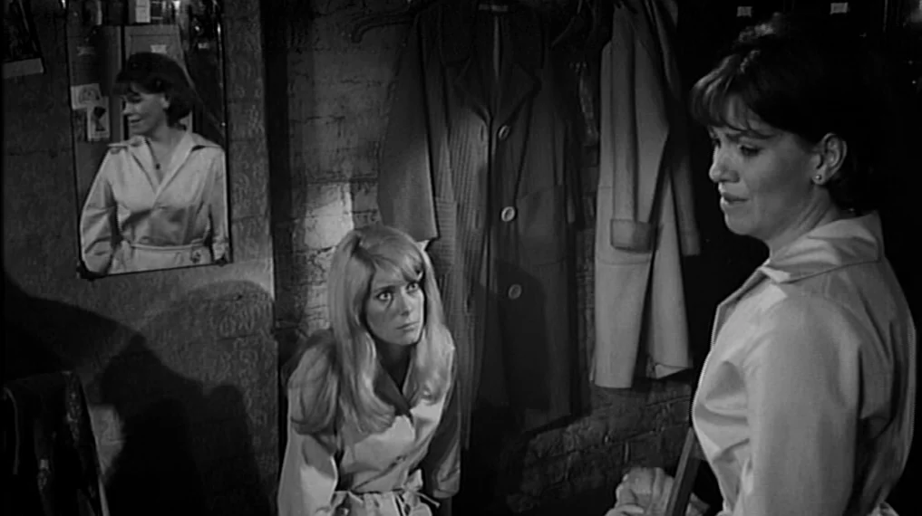 Repulsion (1965) movie still: medium — A cramped dressing room features exposed brick walls and hanging garments on wooden hangers. One woman sits…