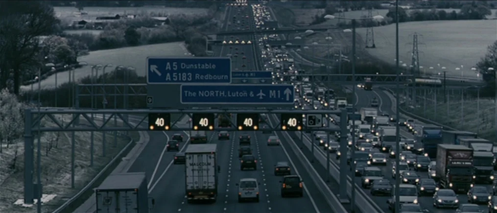 Another Year (2010) movie still: wide — A British motorway scene shows heavy traffic under grey, overcast weather conditions. Numerous cars and…