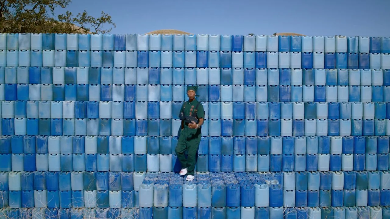 Black is King (2020) movie still: fullbody — A man stands centered before a towering, expansive wall made of stacked blue containers. The…