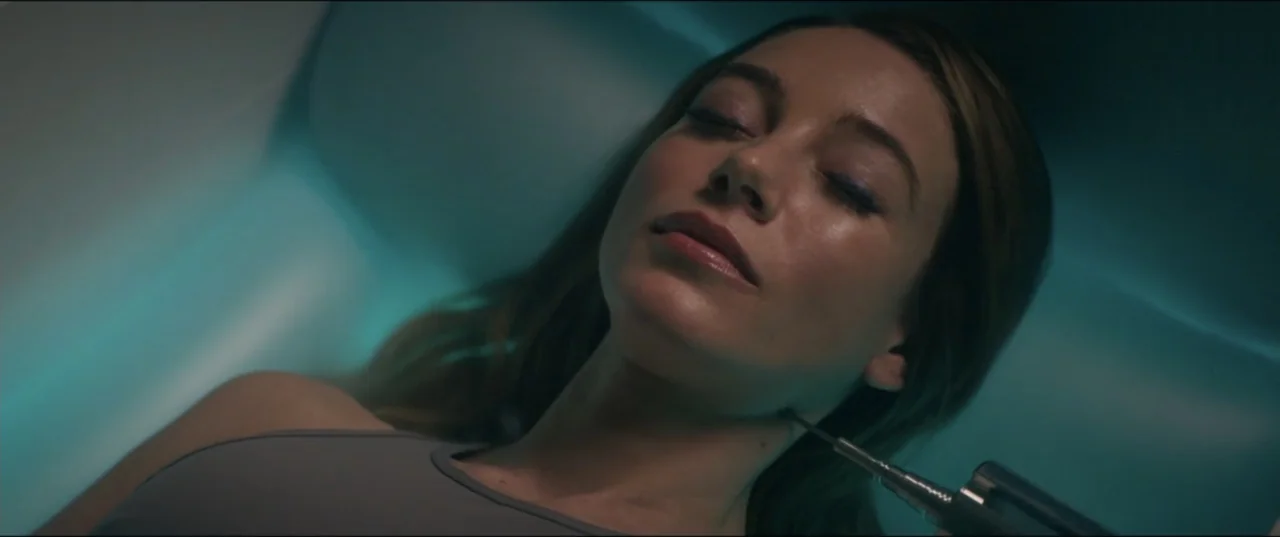 Utopia (2013) movie still: closeup — A futuristic medical pod illuminated by a diffuse cyan light source. A woman with closed eyes lies still as a…