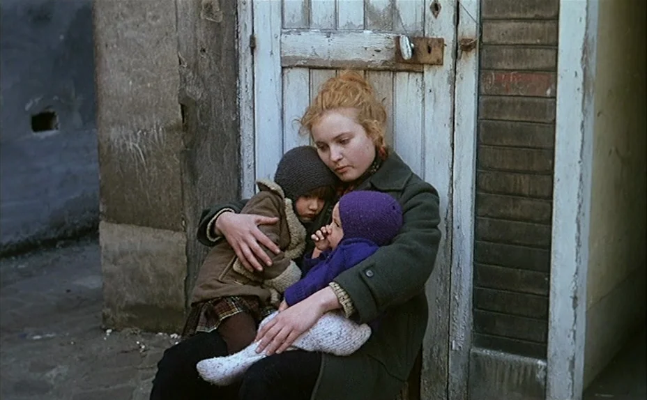 L’une Chante, l’Autre Pas (1977) movie still: medium — Three people sit on the ground against a weathered white wooden door in an alley. The woman…