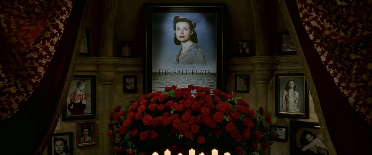 V for Vendetta (2006) movie still: medium — A stone alcove features a large framed portrait of a woman and numerous smaller photos. A dense…
