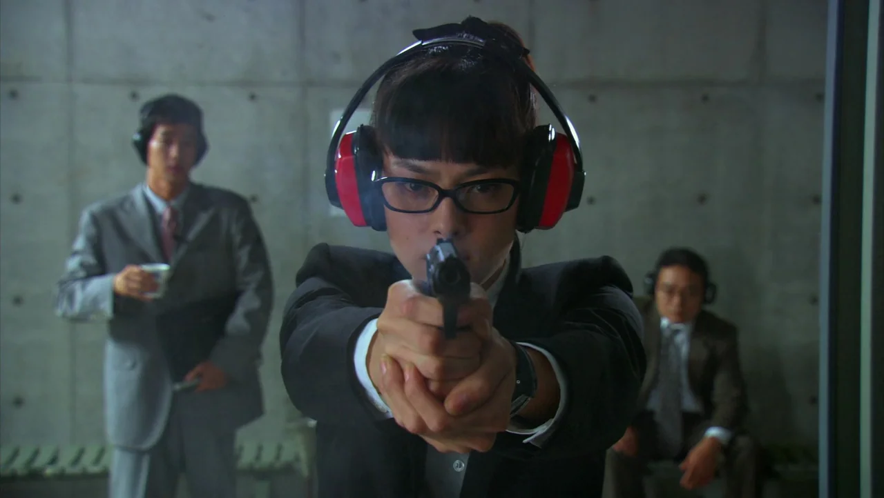 Cutie Honey (2004) movie still: medium — An indoor shooting range features bare gray concrete block walls with visible tie-rod holes. A woman in a…