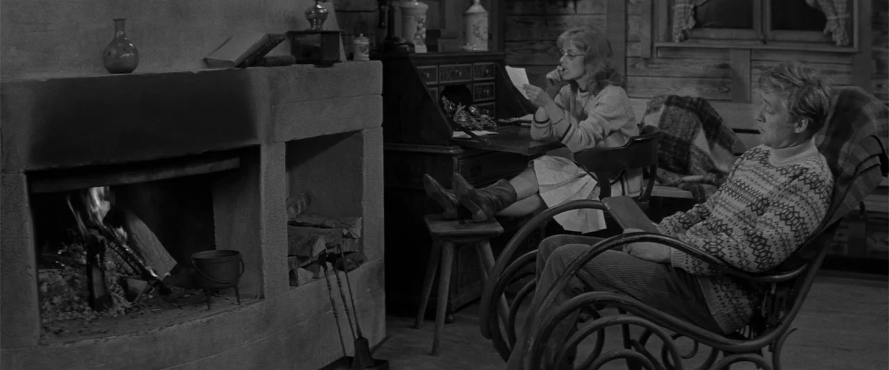 Jules and Jim (1962) movie still: medium — A rustic cabin interior features a stone fireplace and wooden walls at night. A woman reads at a desk…