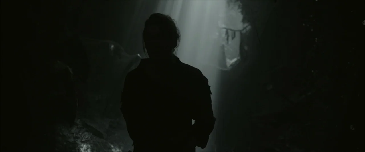 Monster Hunter (2020) movie still: medium — A dark, rocky cavern interior is pierced by a single shaft of natural light. A solitary figure stands…