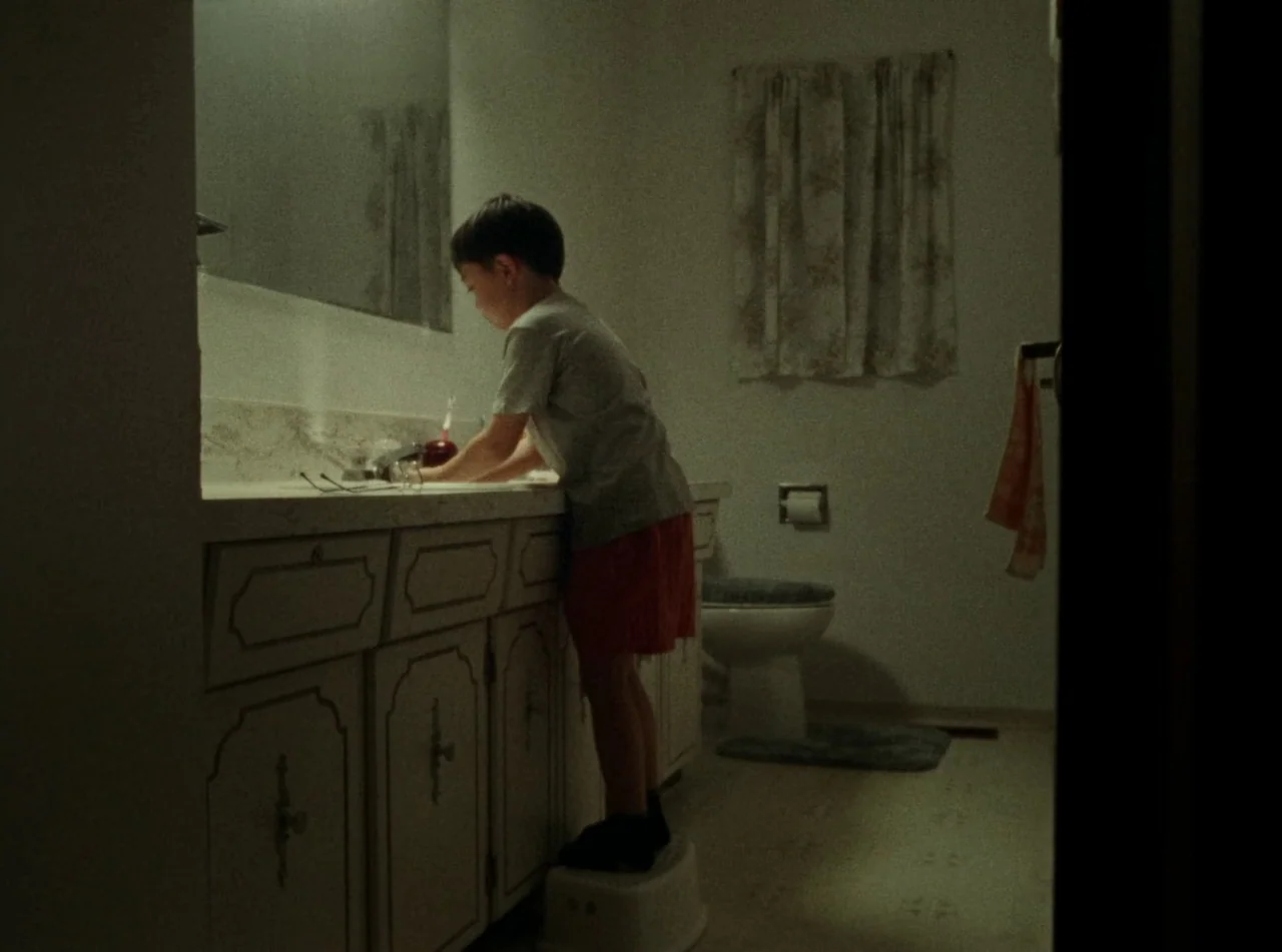 Riceboy Sleeps (2022) movie still: medium — A residential bathroom with patterned curtains and a white cabinet vanity under a mirror. A young boy in…