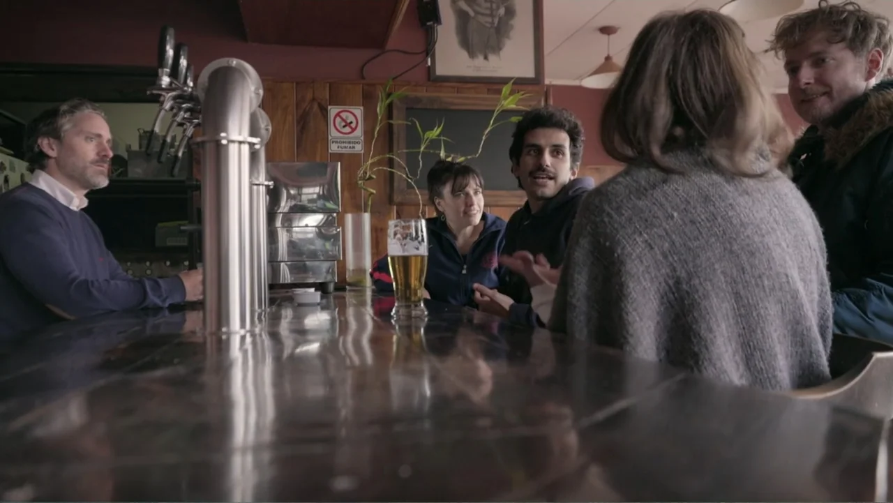 Saturdays Disorders (2022) movie still: medium — A dimly lit bar with dark wood paneling and a long metal counter. Five people sit together talking…