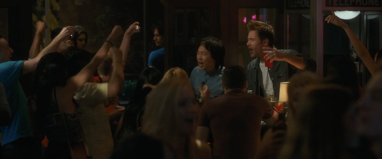 Life of the Party (2018) movie still: medium — A dimly lit bar interior filled with a small group of young people cheering. A man and a woman in the…