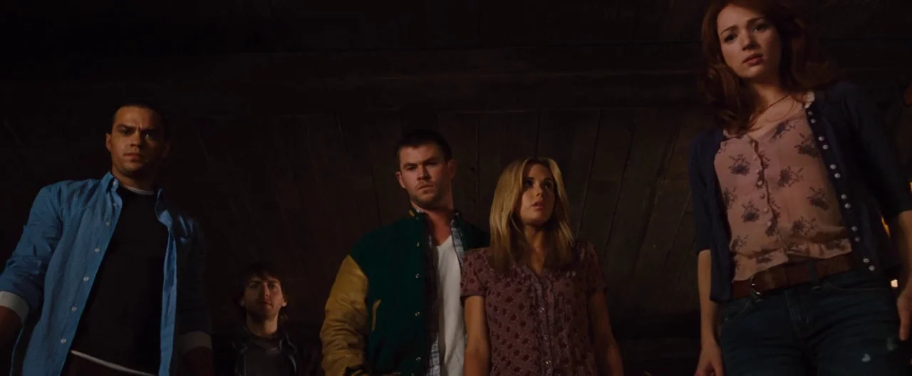 The Cabin In The Woods (2012) movie still: medium — A dimly lit wooden basement with low ceiling beams and shadowed surroundings. Five young adults…