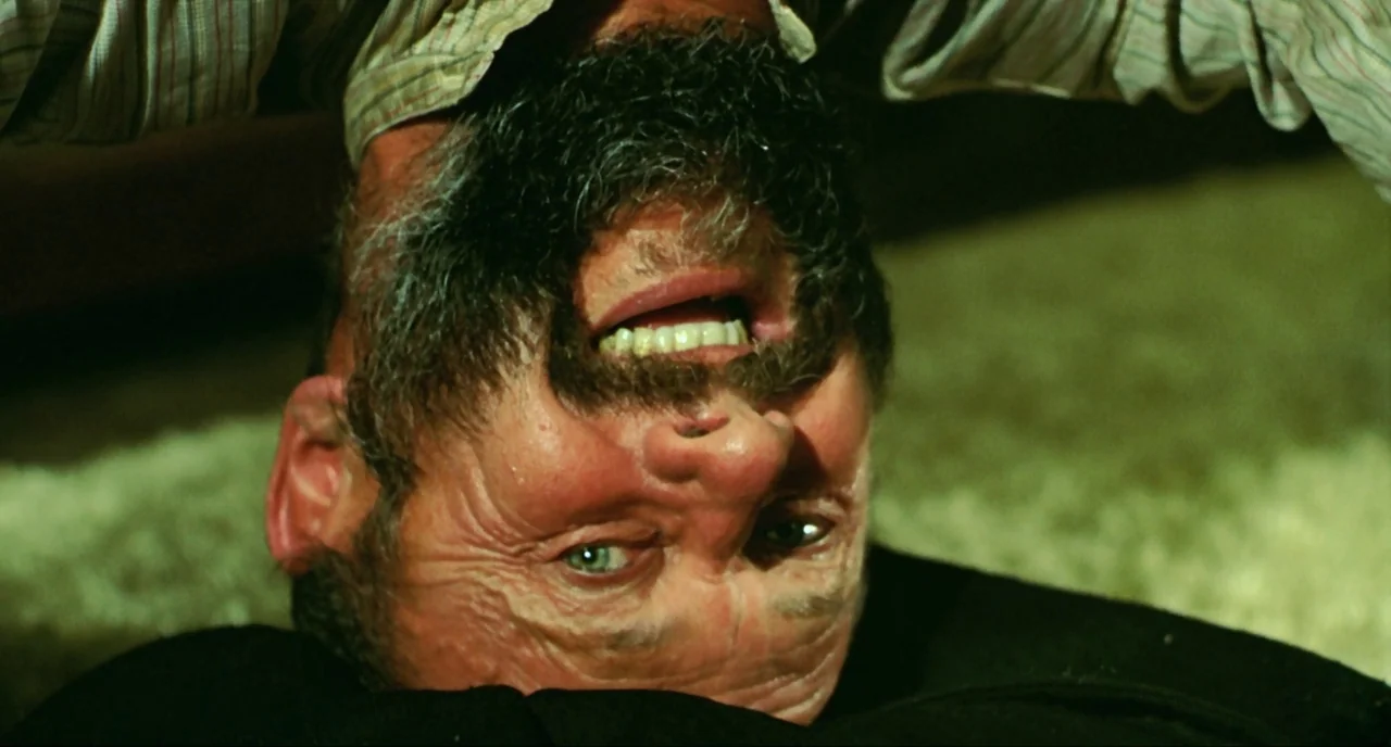 Wake In Fright (1971) movie still: closeup — A man lies inverted on a textured beige rug in a dimly lit room. The man has a thick beard and his…