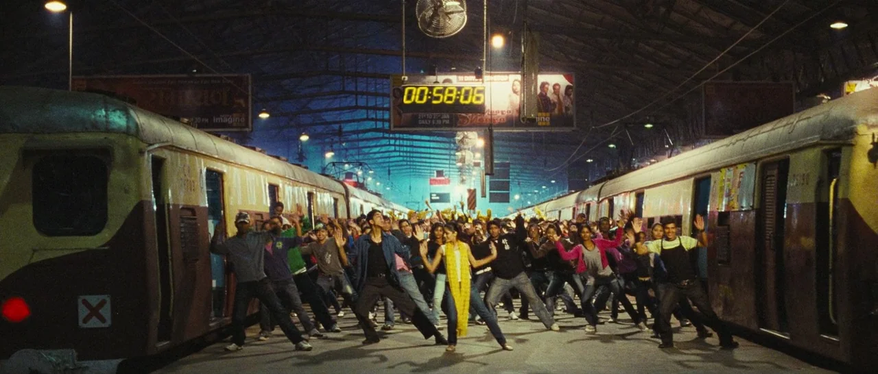 Slumdog Millionaire (2008) movie still: wide — A crowded train station platform at night with two trains parked on either side. Many people are…