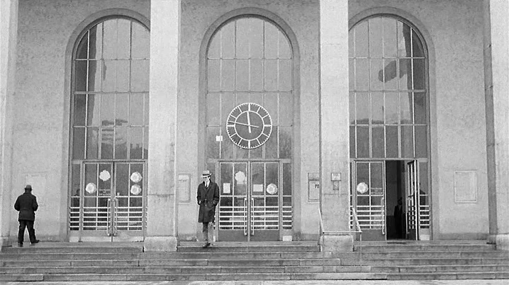 Love Is Colder Than Death (1969) movie still: wide — A stone building with large arched windows and a central clock above the doors. Two men in…