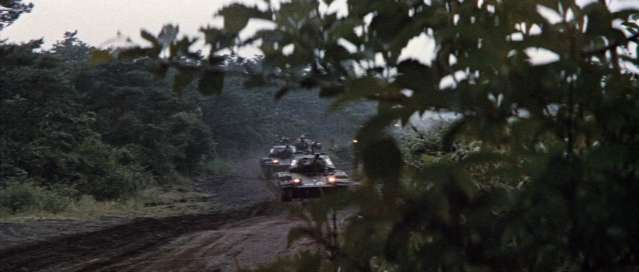 Godzilla Vs Megalon (1973) movie still: wide — A column of tanks travels along a muddy road flanked by dense forest trees. The view is partially…