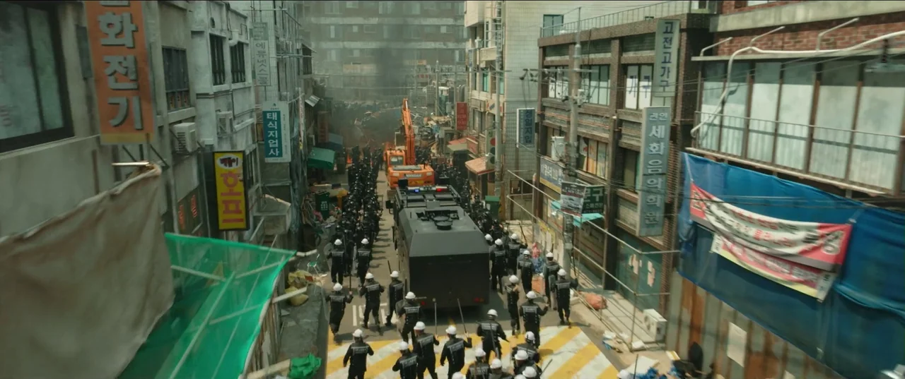 Psychokinesis (2018) movie still: wide — A dense urban street flanked by brick buildings and numerous signage markers in Korean. A column of police…