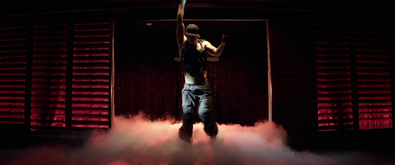 Magic Mike (2012) movie still: fullbody — A dimly lit performance area features red backlighting and dense stage fog. A male dancer jumps mid-air in…