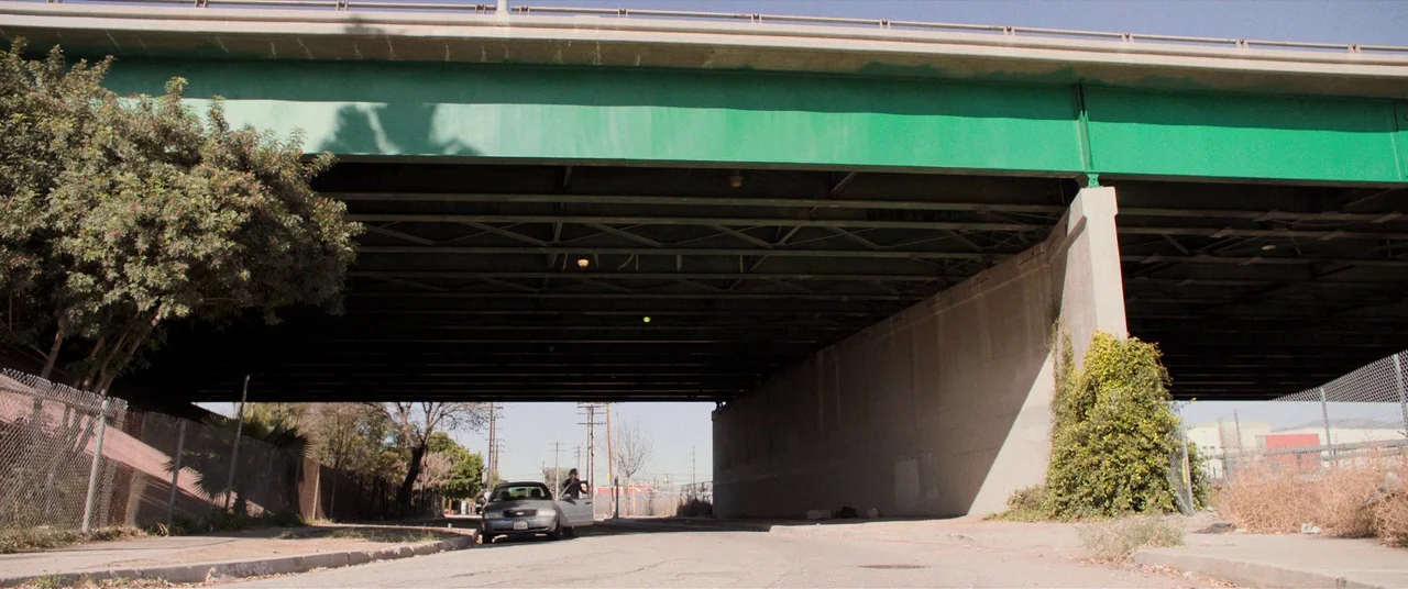 Destroyer (2018) movie still: wide — An asphalt road runs underneath a massive concrete highway bridge with green painted steel beams. A single…