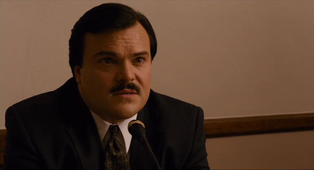 Bernie (2011) movie still: medium — A man in a suit sits before a microphone in a sparse, wood-paneled space. He leans toward the microphone to speak.