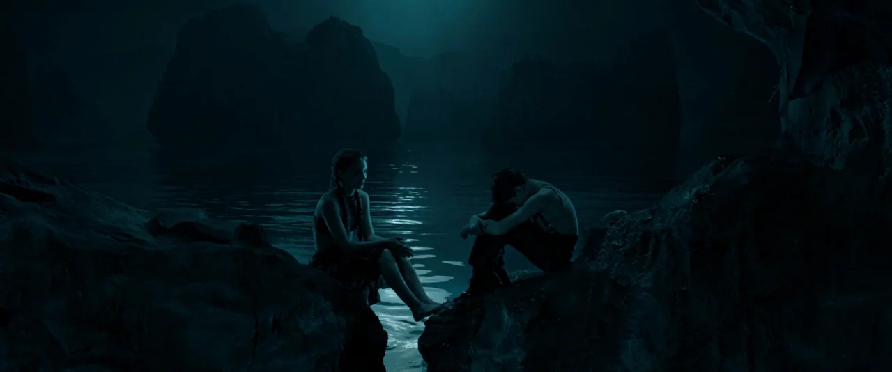 Pan (2015) movie still: wide — A dark rocky shoreline by a sea with jagged karst-like formations in the distance. Two young people sit on separate…