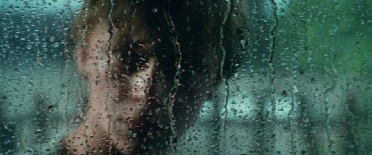 Adoration (2019) movie still: closeup — A close-up view looking through a rain-streaked window pane at a blurred face. A person rests their face…