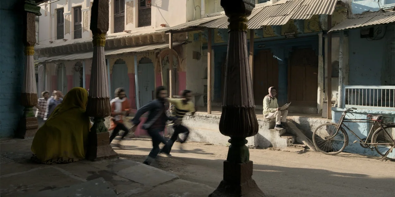 Dry Day (2023) movie still: wide — A dusty Indian street is lined with weathered buildings featuring ornate wooden pillars and balconies. An older…