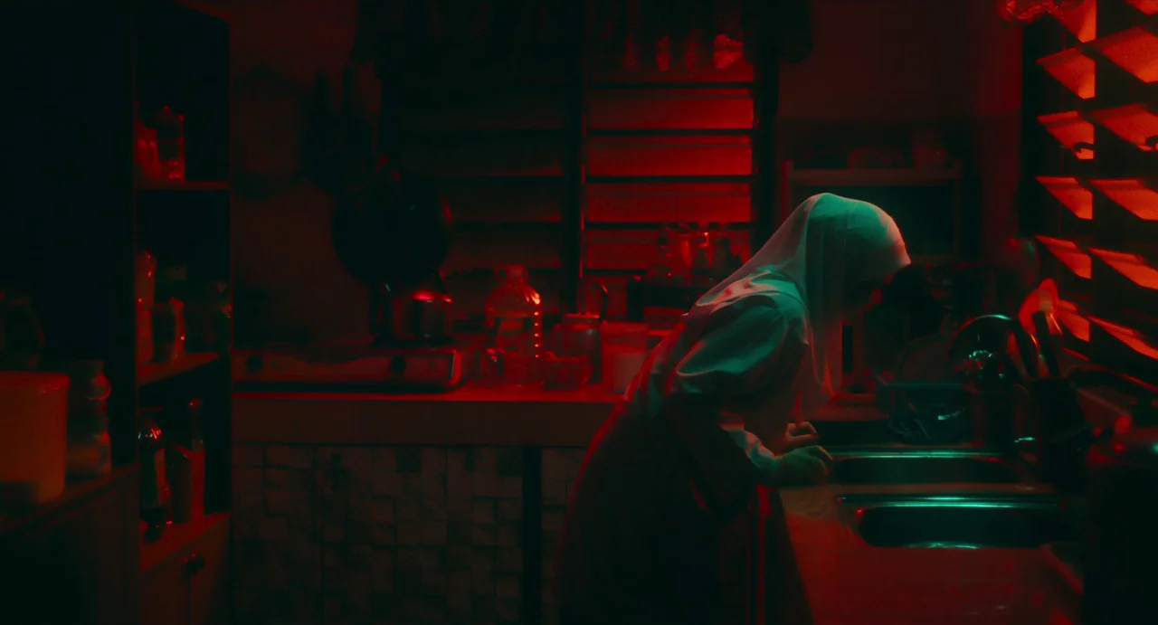 Tiger Stripes (2023) movie still: medium — A dark, cramped space filled with everyday cooking items is washed in red light. A woman wearing a hijab…