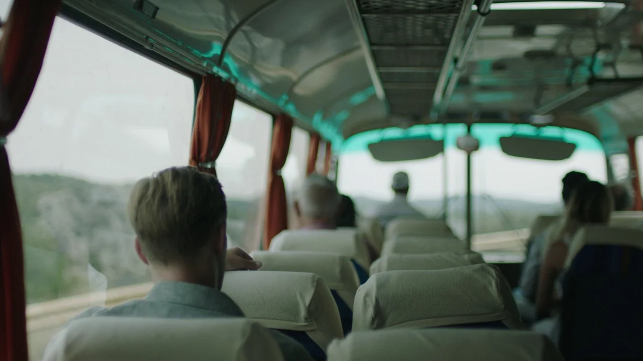 Infinity Pool (2023) movie still: medium — A bus interior with rows of padded seats and orange curtains near the windows. Several passengers sit in…