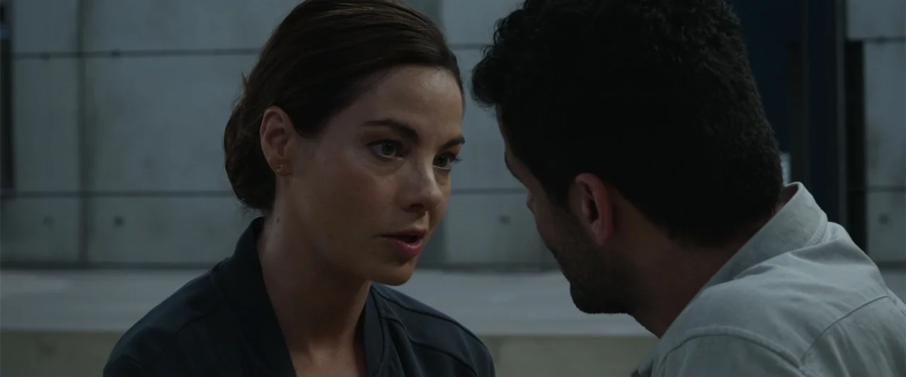Black Site (2022) movie still: medium — A modern building corridor features grey panel walls and clean, minimalist architecture. A woman stands…