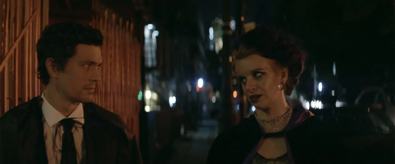 Mennonite's Bite (2023) movie still: medium — Two individuals stand on a dark city sidewalk at night with blurred lights behind them. They wear dark…