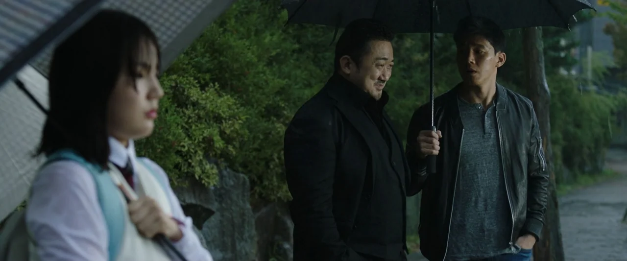 The Gangster, the Cop, the Devil (2019) movie still: medium — Three individuals stand under umbrellas on a paved path lined with dense green…