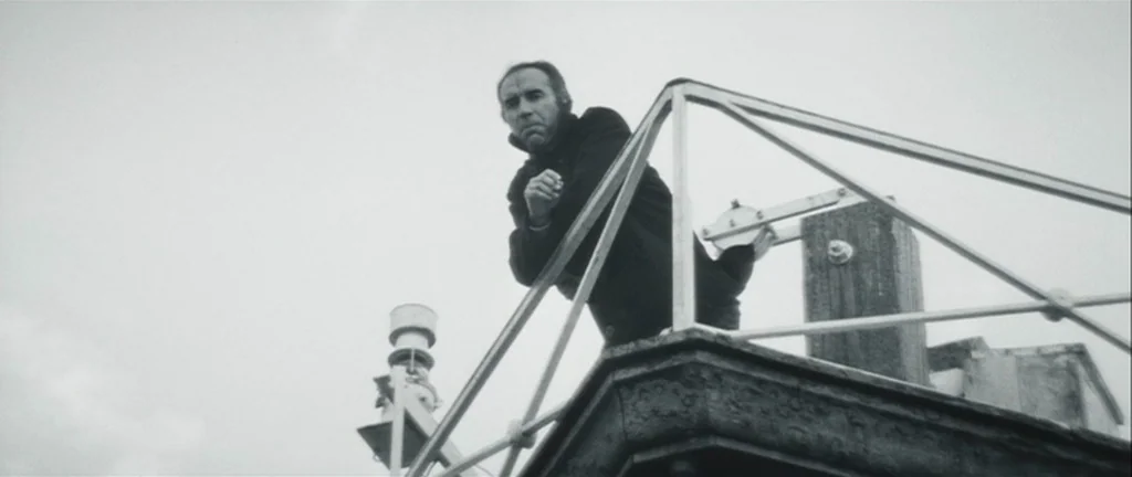Les Créatures (1966) movie still: medium — A man stands on the metal observation deck of a boat under an overcast sky. He leans forward with his…