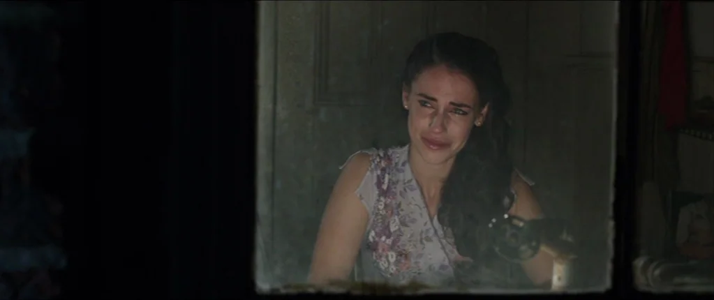 Abattoir (2016) movie still: medium — A dark, dimly lit room is viewed through a glass window pane. A woman sits behind the glass looking out with…